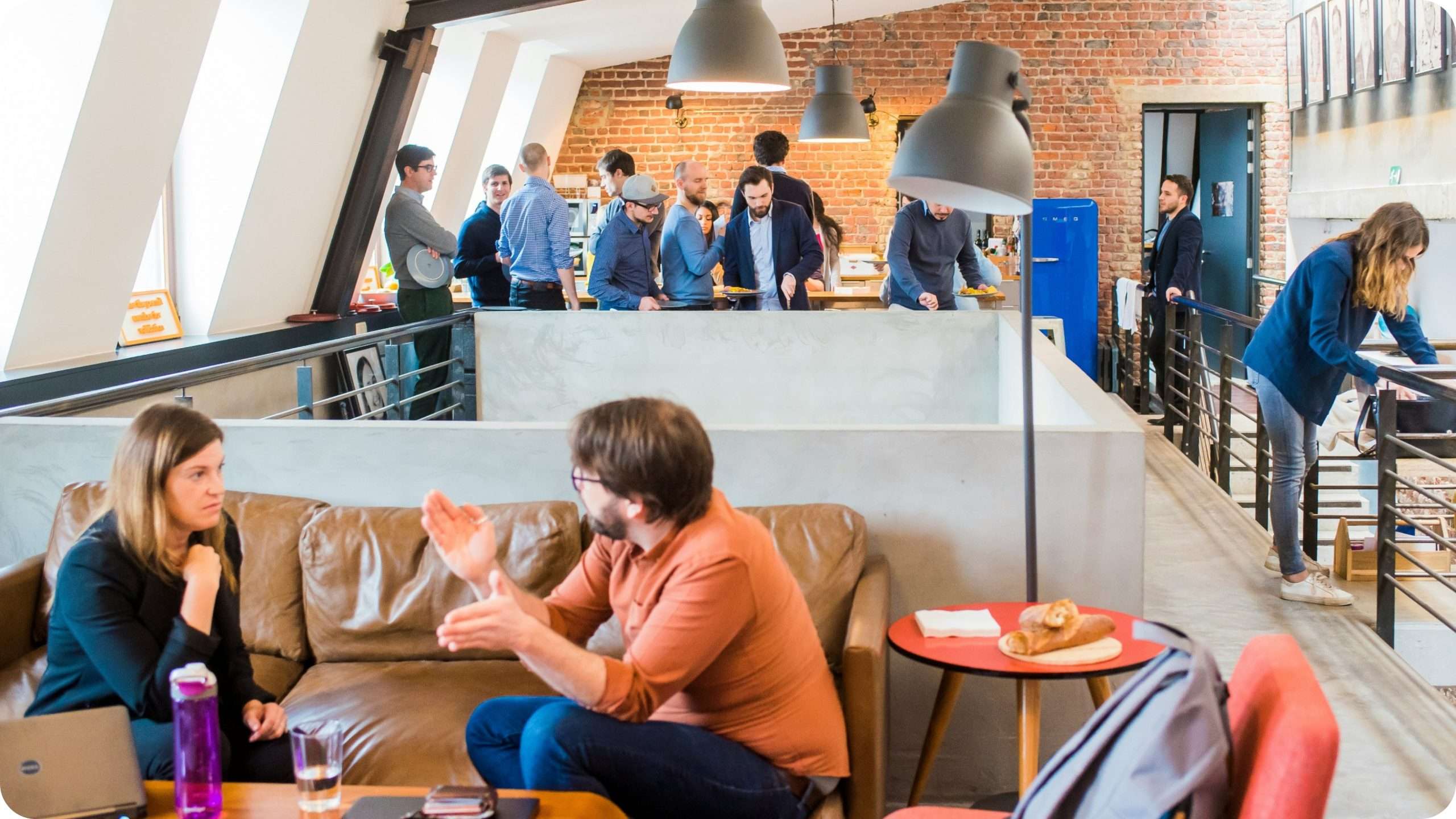 Symbolbild einer offen gestalteten Büro-Küche im Loft-Stil. Im Hintergrund bedienen sich einige Mitarbeiter an einem aufgebauten Buffet, während im Vordergrund zwei Personen in einer Sofa-Ecke sitzen und angeregt zu reden scheinen. Vor Ihnen steht ein aufgeklappter Laptop.