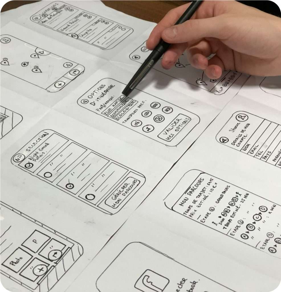 Darstellung mehrerer Wireframes einer Smartphone Anwendung, die auf Papier gezeichnet sind und auf einem Tisch liegen. Eine Hand, die aus der rechten oberen Bildecke kommt zeichnet etwas ein.