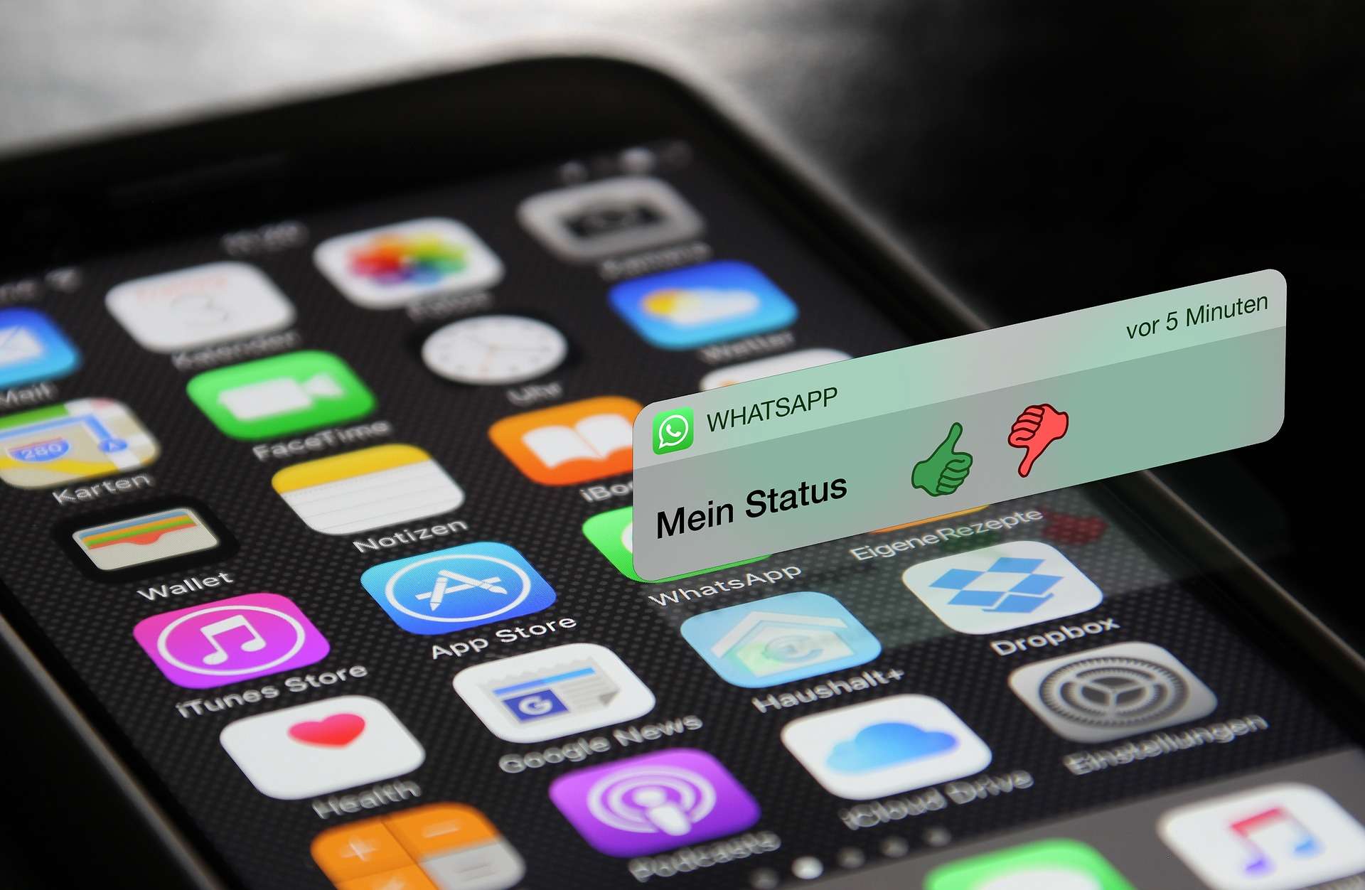 Nahaufnahme eines iPhones, auf dem verschiedene Apps wie FaceTime, Wallet, iTunes Store, App Store, WhatsApp und andere zu sehen sind. Eine WhatsApp-Benachrichtigung mit dem Text 'Mein Status' und Daumen hoch und Daumen runter Symbolen erscheint über dem Bildschirm.